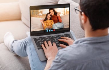 Bir adam kanepede oturuyor, günlük kıyafetler ve gözlük takıyor, karısıyla ve kızıyla görüntülü konuşma yapmak için laptop kullanıyor. Kadın ve kız birlikte oturuyor, adama el sallıyorlar..