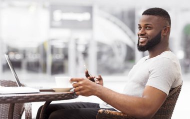 Kafenin dışındaki bir masada oturan bir adam yan tarafa bakıp telefonunu kullanırken gülümsüyor..