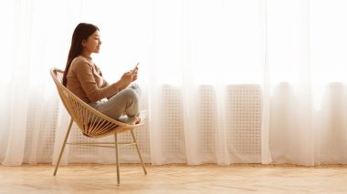 Hasır koltukta oturan genç bir kadın telefonunu kullanıyor. Perdeleri olan bir pencerenin yanında, yumuşak ışığın odayı doldurmasına izin veriyor..