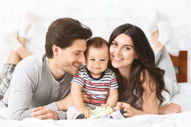 Bu resim, üç kişilik mutlu bir aileyi yatakta yatarken gösteriyor. Ebeveynler gülümsüyor ve ellerinde oyuncakla yatakta oturan küçük çocuklarına bakıyorlar..