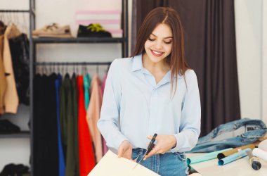 Moda mağazasında tasarım malzemelerini sınıflandıran neşeli genç bir kadın. Arka planda renkli kumaşlar var. Kumaş kesiyor.