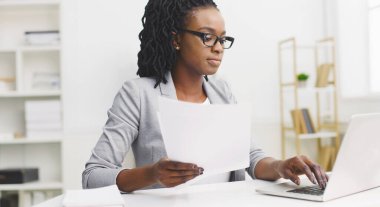 Odaklanmış siyah bir kadın özenle ofis masasında evrak işleriyle uğraşıyor. Ortam profesyonel ve düzenli. Düzenli bir çalışma alanı, modern bir dekor ve parlak bir atmosfer var..