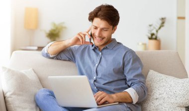 Bir adam kanepede oturuyor, dizüstü bilgisayar kullanıyor ve telefonuyla konuşuyor. Evden çalışıyor gibi görünüyor..