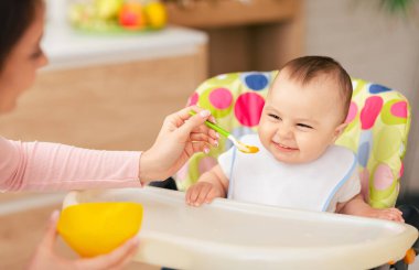 Bir erkek bebek yüksek bir sandalyede oturuyor ve mutfakta annesi tarafından besleniyor. Atmosfer sıcak ve rahatlatıcı. Sevgi dolu ve besleyici bir yemek zamanı..