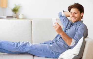 Kot pantolonlu ve mavi gömlekli bir adam beyaz koltukta telefon ve kulaklıklarla rahatlar, rahat bir ifadeyle geriye yaslanır..