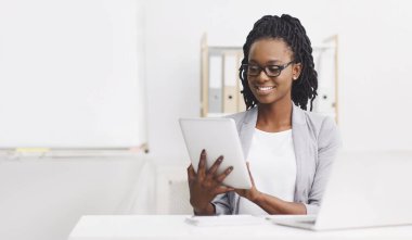 Resmi kıyafet giyen, gözlüklü olumlu genç bayan ofiste dijital tablet kullanarak dizüstü bilgisayarla masada oturuyor.