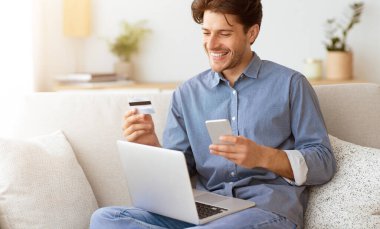 Kanepede oturan bir adam elinde kredi kartıyla elinde bir dizüstü bilgisayarla telefonuna bakıp gülümsüyor ve online ödeme yaptığını ima ediyor..