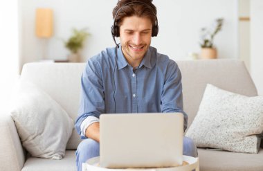 Kulaklık takan bir adam evinde kanepede oturur, gülümser ve dizüstü bilgisayarla çalışır..