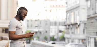 Kulaklık ve güneş gözlüğü takan bir adam şehirde yürürken telefonunu kullanıyor..