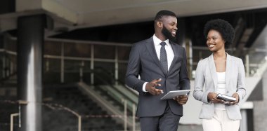 Birlikte açık havada yürüyen iki gülümseyen Afrikalı iş arkadaşları, elinde dijital tablet tutan adam ve not defteri tutan bir kadın. Boş alan