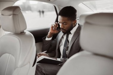 Takım elbiseli siyah bir adam arabanın arka koltuğunda oturur, bir telefon görüşmesi sırasında not defterine not alır. Odaklanmış ve ciddi, muhtemelen önemli iş meselelerini tartışıyor..