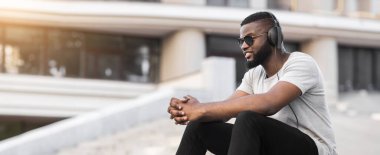 Kulaklık takan genç bir adam şehir ortamında bir basamakta oturuyor. O, güneş gözlüğü ve beyaz bir gömlek takıyor ve yana bakıyor..