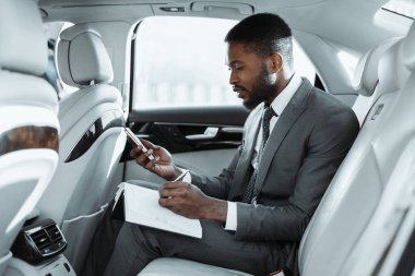 Takım elbiseli siyah bir adam arabanın arka koltuğunda oturur ve not defterine notlar yazarken akıllı telefonu üzerinde çalışır. İşine odaklanmış ve meşgul görünüyor.