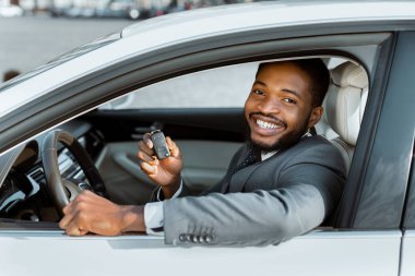 Takım elbiseli siyah adam yeni bir arabanın sürücü koltuğunda otururken gülümsüyor ve bir araba anahtarı tutuyor. Aracın sahibi olduğu için mutlu ve heyecanlı görünüyor..
