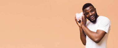 Sakallı, kulağına pembe domuz kumbarası tutarak gülen Afro-Amerikalı bir adam, görünüşe göre dinliyor. Beyaz bir tişört giyiyor ve yüzünde dostane bir ifade var.