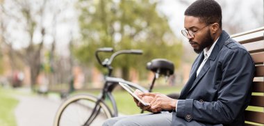 Afrikalı Amerikalı bir adam parktaki bankta dijital tablet üzerinde çalışıyor. Ortalık sakin ve yeşildir. Doğada iş ve eğlence karışımını yansıtır..