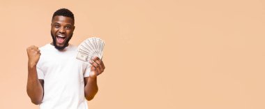 Geniş gülümseyen ve yumruğunu kaldırmış siyah bir adam elinde bir para fanatiği tutuyor. Beyaz bir tişört giyiyor ve açık şeftali rengi bir arka plan üzerinde duruyor.