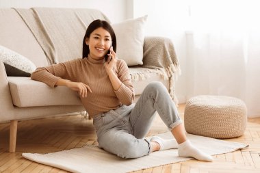 Oturma odasında halıya oturmuş cep telefonuyla gülümseyen genç bir kadın..