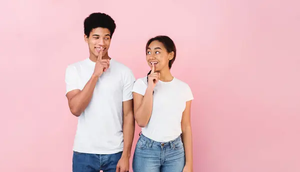 stock image Shhh, its our secret. Teen couple holding fingers on lips and looking at each other, pink studio background