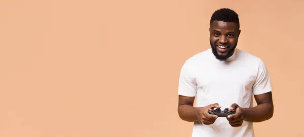 stock image A smiling black man is shown holding a video game controller in front of a peach-colored background. The man appears to be enjoying himself and is likely immersed in a video game, copy space