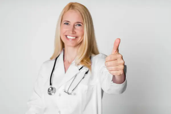 stock image A blonde woman doctor wearing a white coat and a stethoscope gives a thumbs up gesture while smiling. She is photographed against a white background.