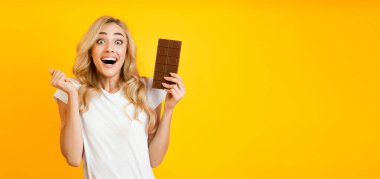 Uzun sarı saçlı genç bir kadın sağ elinde çikolata tutuyor ve ağzı açık heyecanlı bir şekilde gülümsüyor. O, parlak sarı bir arka planda duruyor..