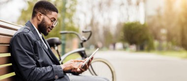 Afrikalı bir Amerikalı işadamı, parktaki bir bankta otururken dijital tablet kullanıyor. Havası sakin, üretken, doğayla iç içe olan bir iş..