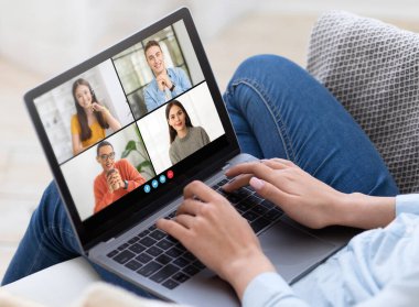 Bir kadın kanepeye oturur ve video konferansı için dizüstü bilgisayar kullanır. Dizüstü bilgisayar ekranında dört katılımcı ayrı video çerçevelerinde gösteriliyor, kırpılmış