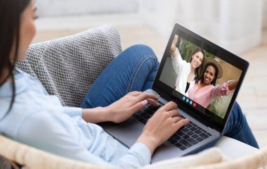 Bir kadın evinde bir kanepede oturuyor, iki arkadaşıyla görüntülü konuşma yapmak için dizüstü bilgisayar kullanıyor. Kadın klavyede yazı yazarken arkadaşlar gülümsüyor ve kameraya el sallıyorlar..