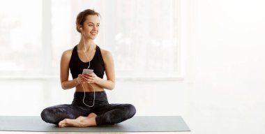 Bir kadın yoga minderine oturmuş kulaklıkla müzik dinlemekle meşgul. Odaklanmış ve rahatlamış görünüyor, muhtemelen farkındalık rutinine müzik de ekliyor..