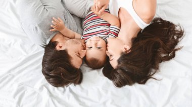 Üç kişilik mutlu bir aile yatakta birlikte yatıyor. Ebeveynler yeni doğan bebeklerini yanaklarından öpüyor. Bebek çizgili bir gömlek giyiyor ve kameraya bakıyor.