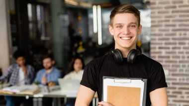 Neşeli genç bir öğrenci elinde bir not panosuyla ön planda duruyor ve kulaklıkları boynunda parlak bir gülümsemeyle.