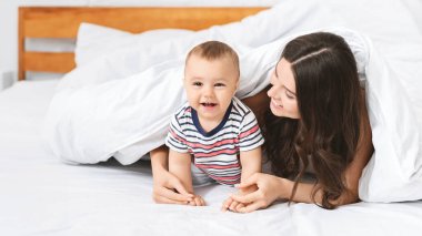 Çizgili gömlek giymiş genç bir bebek annesiyle birlikte beyaz bir yatakta yatarken kameraya doğru sürünüyor. Bebek gülümsüyor ve emeklemekten mutlu görünüyor..
