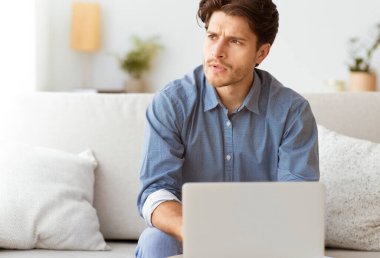 Mavi gömlekli bir adam evinde bir kanepede oturur, bir dizüstü bilgisayarla çalışırken dikkatlice yan tarafa bakar..