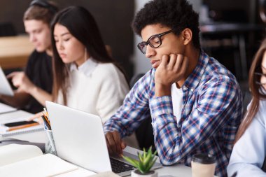 A multiethnic group of focused young adults is sitting at desks in a classroom setting. They appear to be absorbed in their individual tasks, with some using laptops and others taking notes clipart