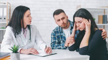 Beyaz önlüklü bir doktor yazı tahtası ve kalemle bir masada oturuyor ve genç bir çiftin endişelerini dikkatle dinliyor. Kadın gözle görülür şekilde üzgün, kafasını elinde tutuyor.