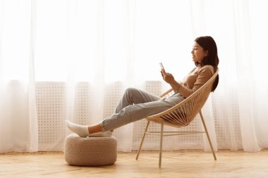 Bir kadın örgü bir sandalyede oturuyor ve telefonuna bakıyor. Ayakları bir sedirde dinleniyor..