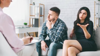 Ofisteki koltukta oturan bir çift terapistle konuşuyor. Kadın eliyle konuşuyor ve işaret ediyor, adam ise dinlerken ve düşünceli düşünürken. Terapist not alıyor..