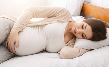 Yorgun ama mutlu hamile bir kadın yatakta yatıyor, yürüyüşten sonra dinleniyor ve karnını okşuyor.