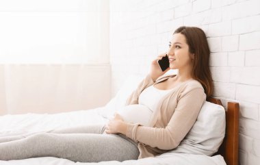 Rahatlamış hamile bir kadın telefonda konuşurken bir yatakta oturuyor. Rahatlamış ve sohbete dalmış görünüyor, modern iletişim ve günlük yaşamın tipik bir anını gösteriyor..