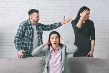 Bir kadın ellerini kulaklarının üzerine koyarak koltukta oturur ve bir erkek kanepenin arkasında duran başka bir kadına bağırır. Kanepenin arkasında eli alnında duran kadın..