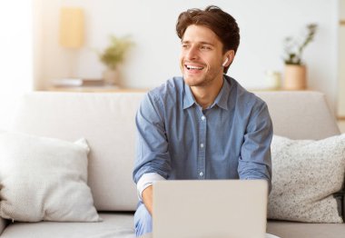 Bir adam kanepede oturuyor, dizüstü bilgisayar kullanırken gülüyor. Mavi düğmeli gömlek ve kulaklık takıyor..