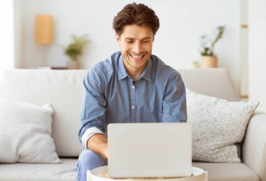 Bir adam kanepeye oturur ve dizüstü bilgisayar kullanırken gülümser..