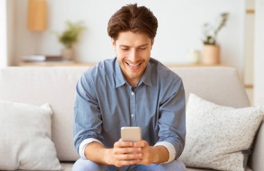 Bir adam koltukta oturuyor, gülümsüyor ve telefonuna bakıyor..