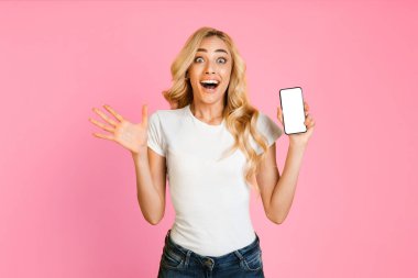 Genç sarışın bir kadın pembe bir arka planın önünde duruyor. Beyaz tişört ve kot pantolon giymişti ve sağ elinde boş bir ekranla akıllı telefonu tutarken çok heyecanlı görünüyordu..