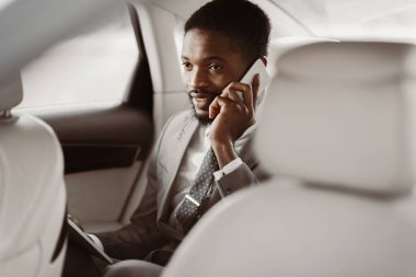 Takım elbiseli Afrikalı Amerikalı bir menajer arabanın arka koltuğunda oturur ve telefonuyla konuşur. Telefona dikkatle bakıyor ve bir sohbete dalmış gibi görünüyor..
