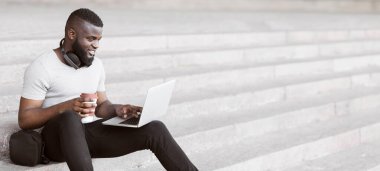 Beyaz tişörtlü bir adam basamaklarda oturuyor, dizüstü bilgisayar kullanıyor ve elinde bir fincan kahve tutuyor..