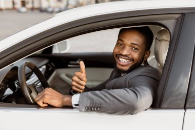 Takım elbiseli Afro-Amerikalı adam gülümseyip, beyaz bir arabanın sürücü koltuğunda otururken başparmağını kaldırıyor..