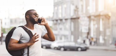 Sırt çantalı genç bir adam şehir merkezinde telefonla konuşurken gülümsüyor..
