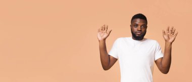 Beyaz tişörtlü, kısa saçlı ve sakallı genç siyah bir adam açık arkaplanın önünde duruyor. O, ciddi bir ifadeyle kameraya bakıyor ve iki elini de onun önünde kaldırdı.